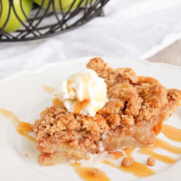 A piece of apple crumble pie on a white plate with ice cream and caramel on top.