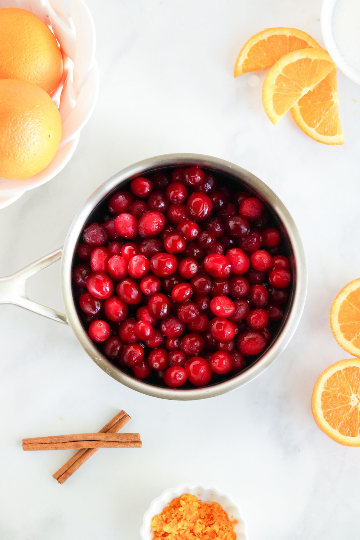 Cranberry Orange Sauce Recipe, a pan of fresh cranberries with whole oranges in the upper left coverner and sliced oranges in the upper right corner.