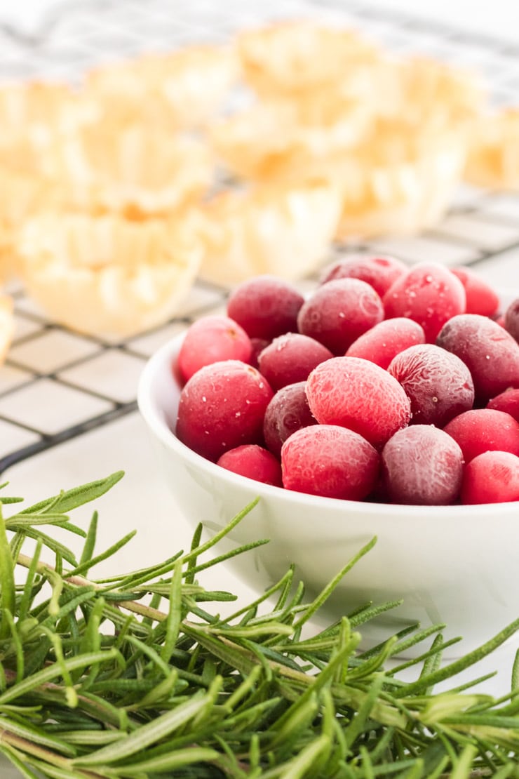 Cranberry Brie Bites- a macro close up image of frozen cranberries, with rosemary in front and pyllo dough in back