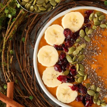 A half bowl shot of a pumpkin smoothie bowl garnished with bananas, cranberries, pumpkin seeds and chia seeds.