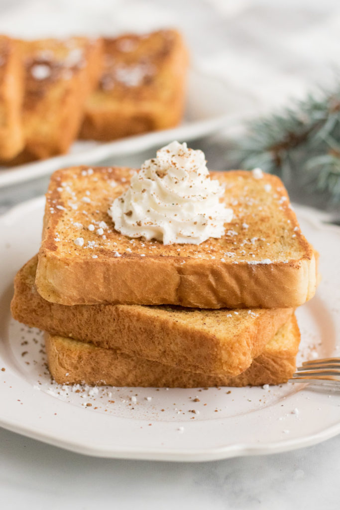 Three pieces of egg nog french toast on a white plate with whipped cream on top. 