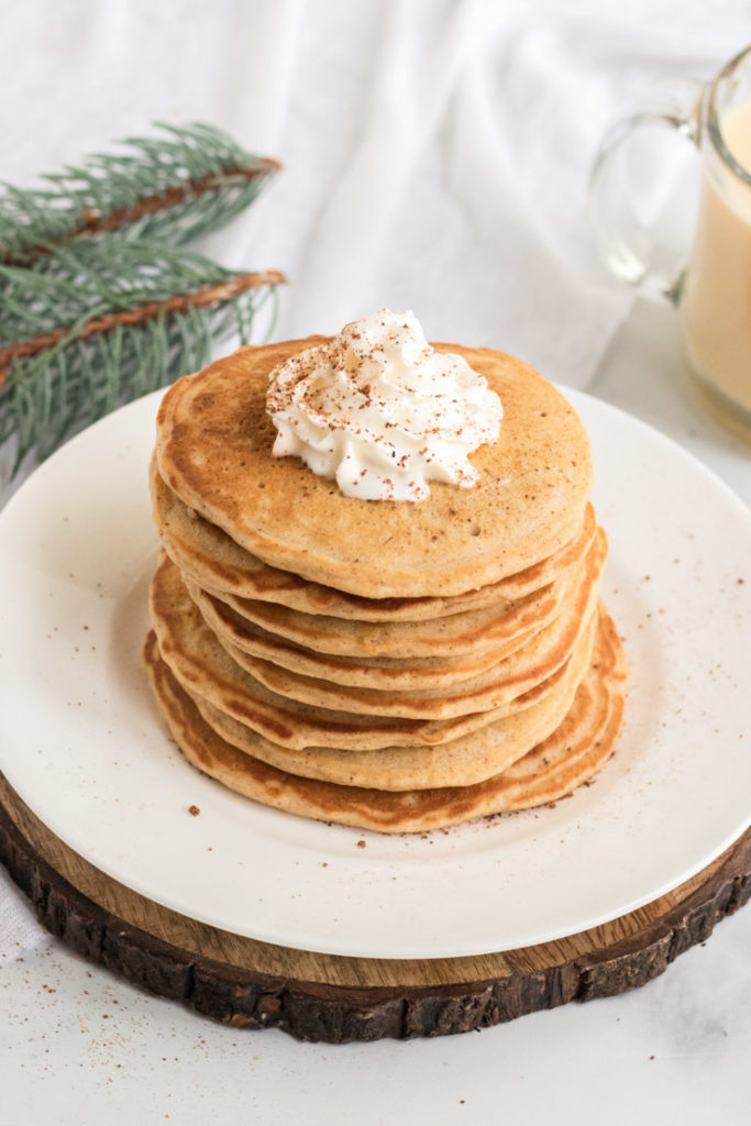 A stack of egg nog pancakes on a white plate on a peice of wood. 