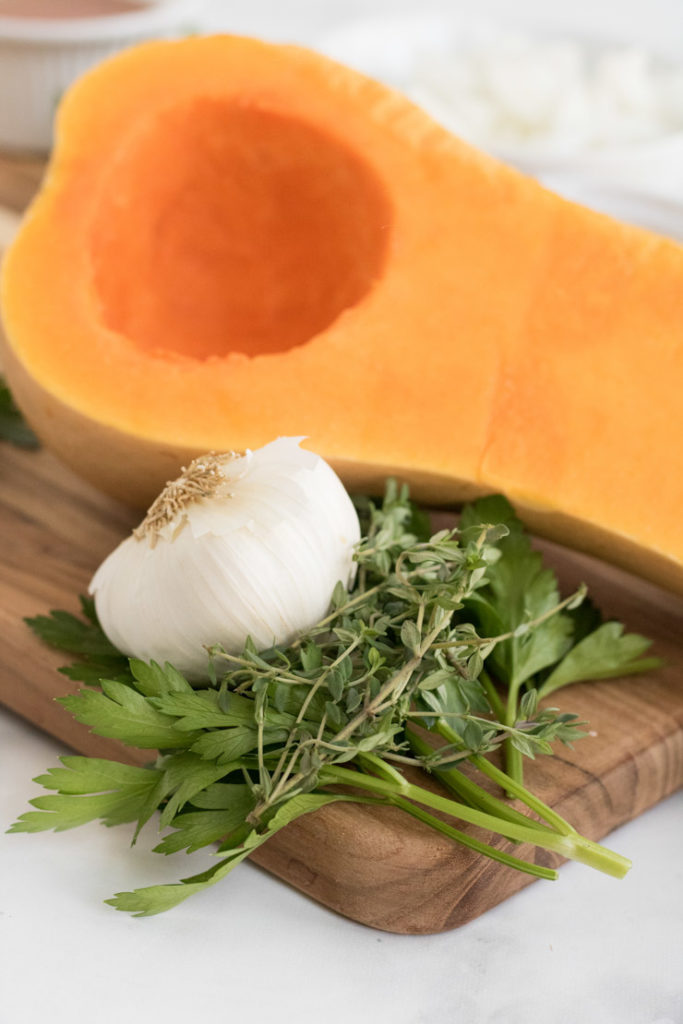 An open faced, raw butternut squash with a head of garlic, parsley and thyme. 