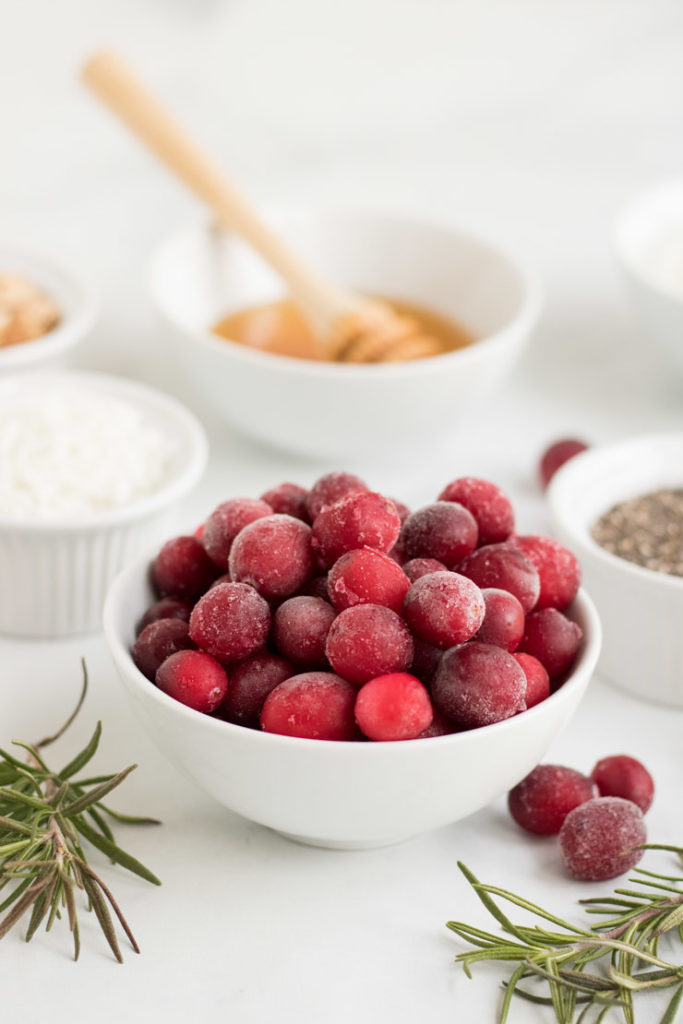 Ingredients for cranberry smoothie bowl. There is a close up of frozen cranberries in a white bowl. In the background there is a bowl of honey, chia and coconut. 