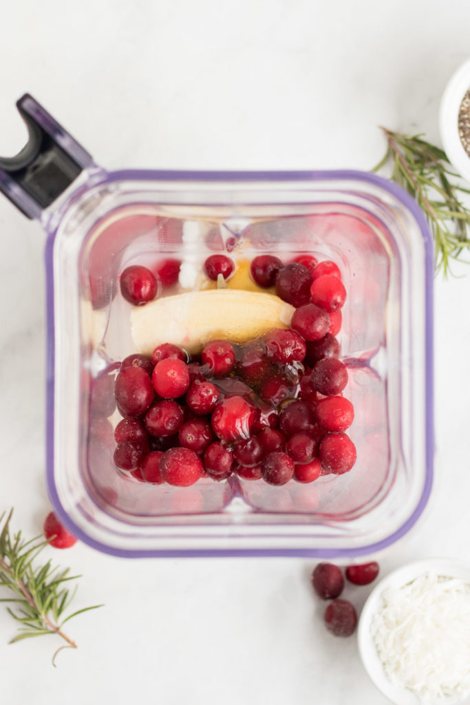 Cranberries, banana, yogurt and honey in a blender. There is rosemary and a few cranberries around it. 