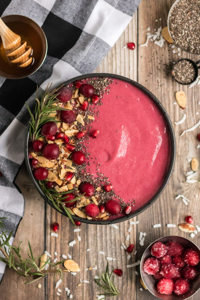 A cranberry smoothie in a black bowl with a wooden background. It is garnished with almonds, cranberries, pomegranates, chia seeds and rosemary.