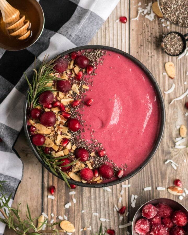 Cranberry smoothie in a black bowl. It is garnished with almonds, cranberries, chia and rosemary.