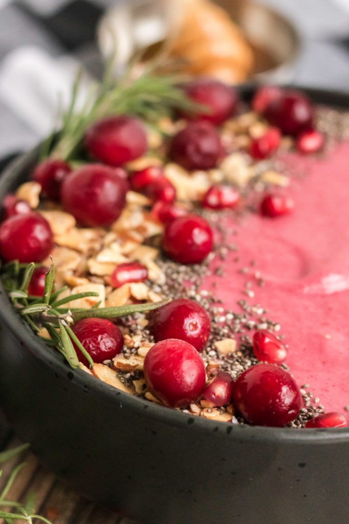 Close up image of a cranberry smoothie bowl with fresh cranberries, chia seeds, almonds and rosemary for garnish. 