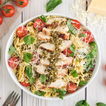 An overhead image of a bowl of chicken pesto pasta salad with basil and tomatoes around it.