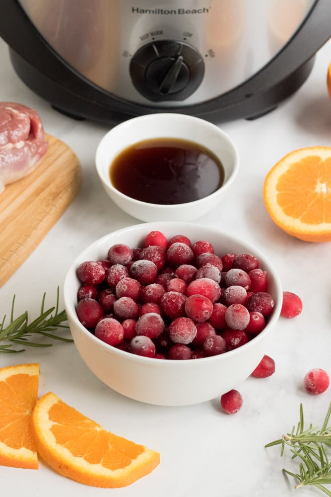 Ingredients for cranberry orange pork .Cranberries in a white bowl, orange slices and maple syrup. 