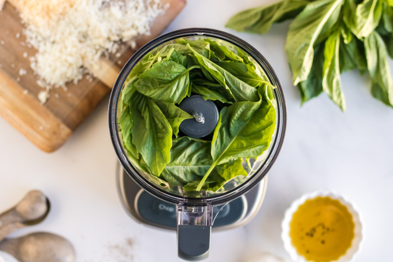 Nut free basil pesto- a photo of basil in a food processor with Parmesan cheese and olive oil around it. 