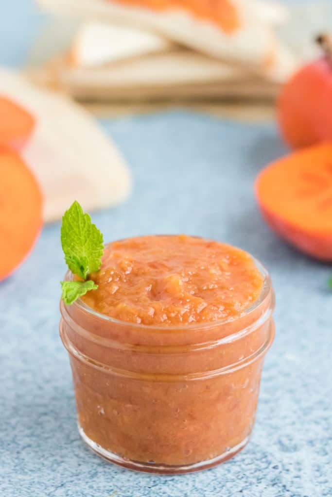 A jar of persimmon jam  with a sprig of mint on a blue background. 