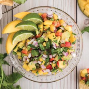 A overhead image of a bowl of freshpico de gallo with mango with limes and mangoes as garnish.