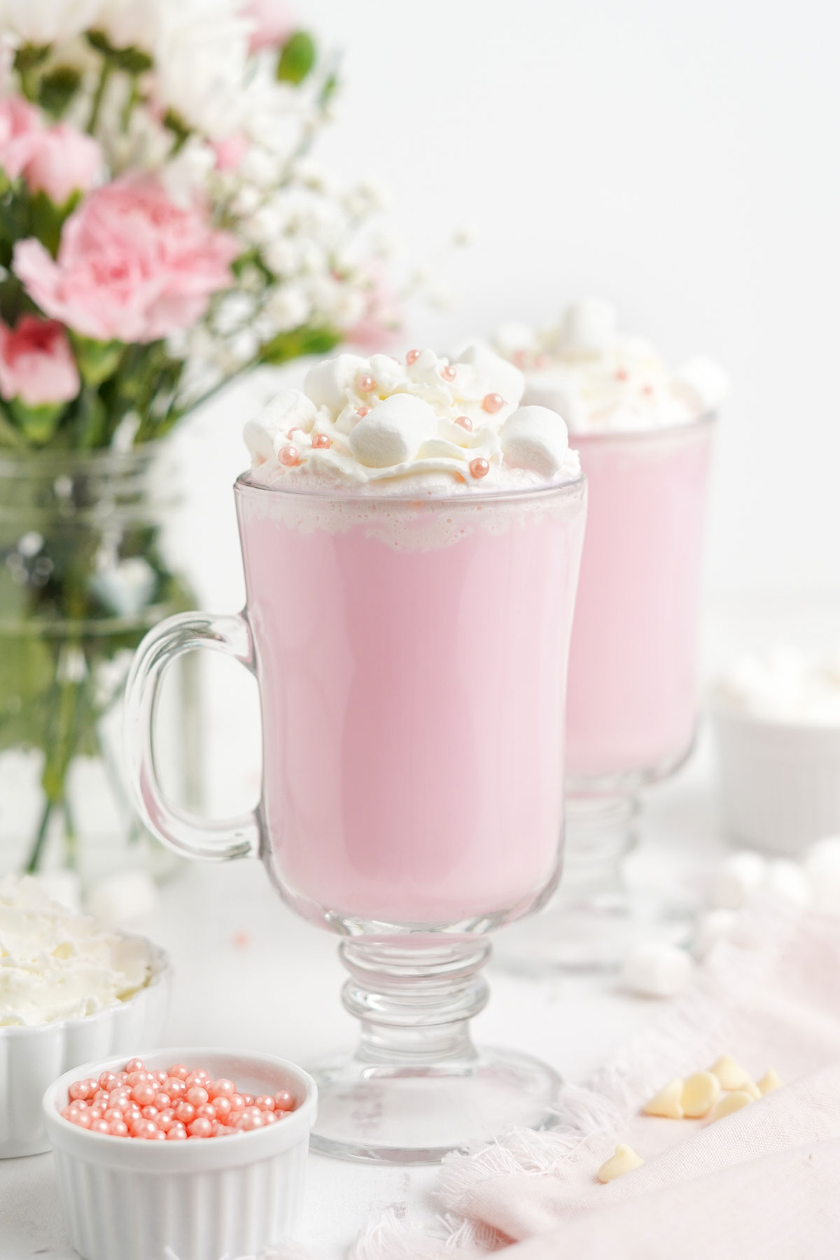 Two glass mugs of pink hot chocolate on the table with pink sprinkles and white chocolate.