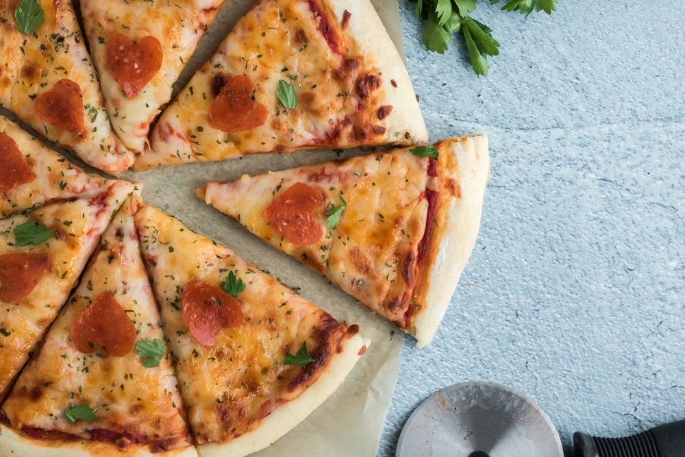 An overhead image of a Valentines Day Pizza. The pizza has pepperoni which is cut out like hearts and is on a blue background. 