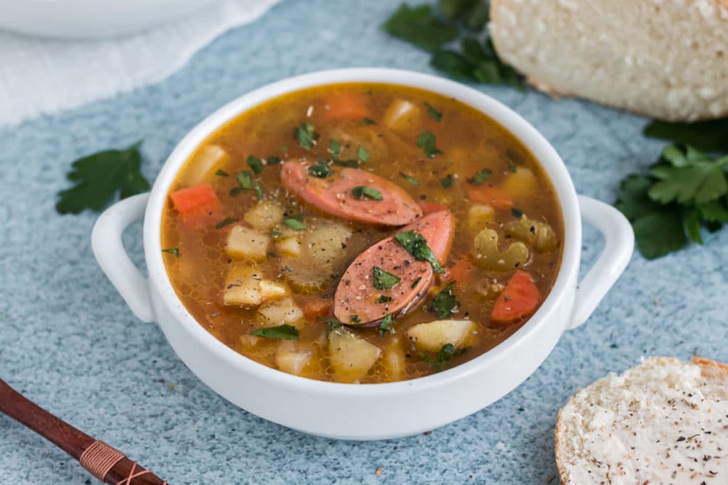 Kartoffelsuppe or German Potato soup in a white bowl with handles on a blue background. The soup has two slices of sausage on cop. 
