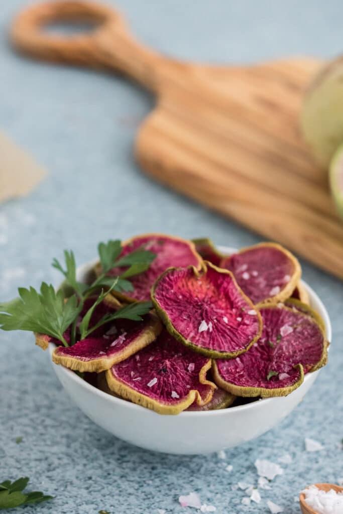 Roasted Watermelon Radish Chips in a light bowl with course salt on a blue background