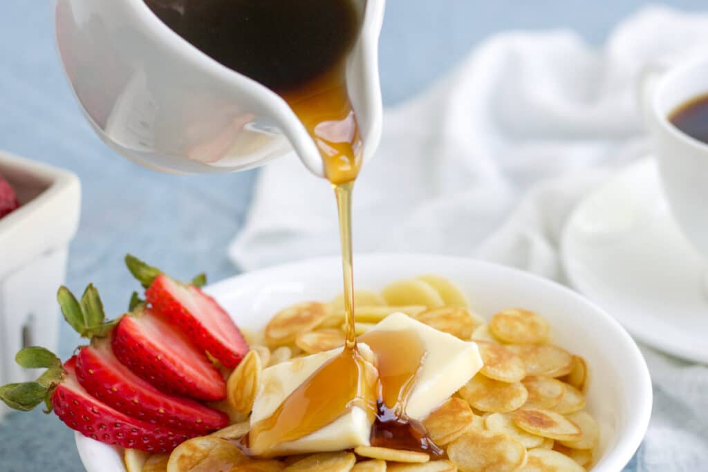 Syrup being poured on mini pancake cereal in a white bowl