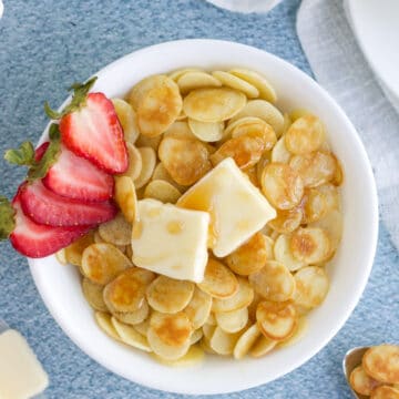 An overhead image of a bowl of pancake cereal in a white bowl with butter and strawberries.