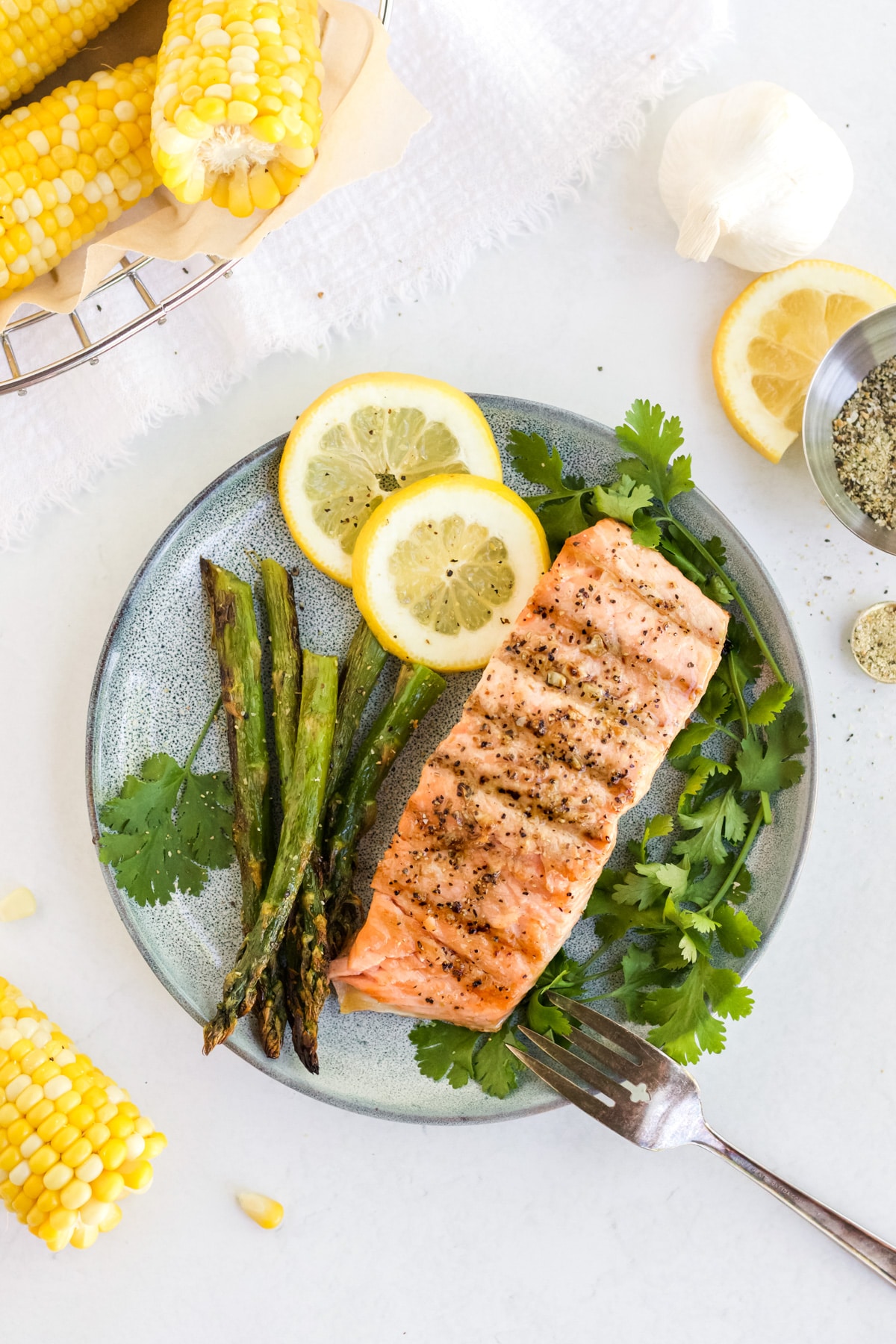 Salmon filet on a plate with herbs, asparagus and lemon, corn on the cob is off in the corner. 