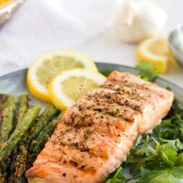 Grilled salmon on a bed of herbs with slices of lemon in the background.