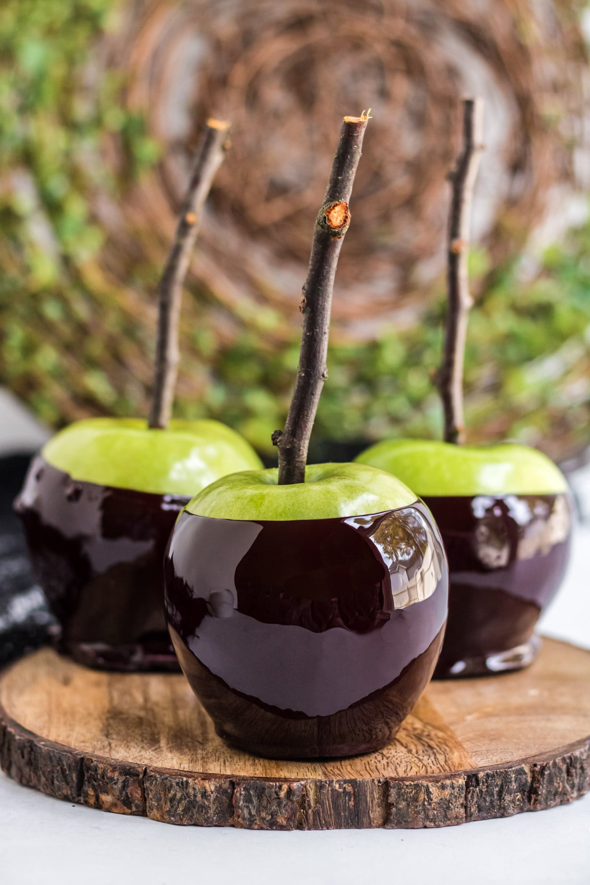 A picture of three black candied apples on a wooden server with sticks in the top. 