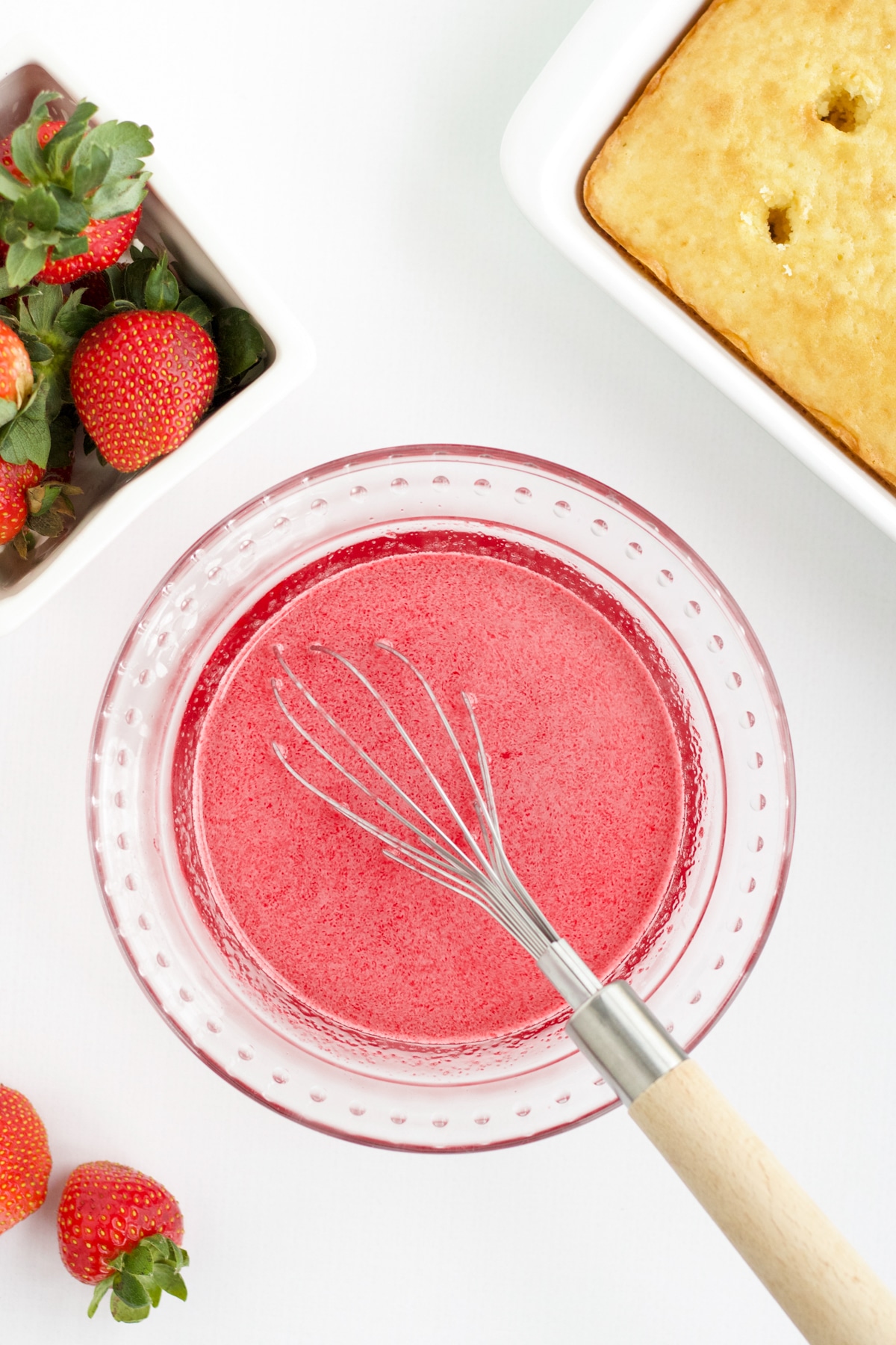 bowl of strawberry jello mixture and a whisk with fresh strawberries and a cake in the background