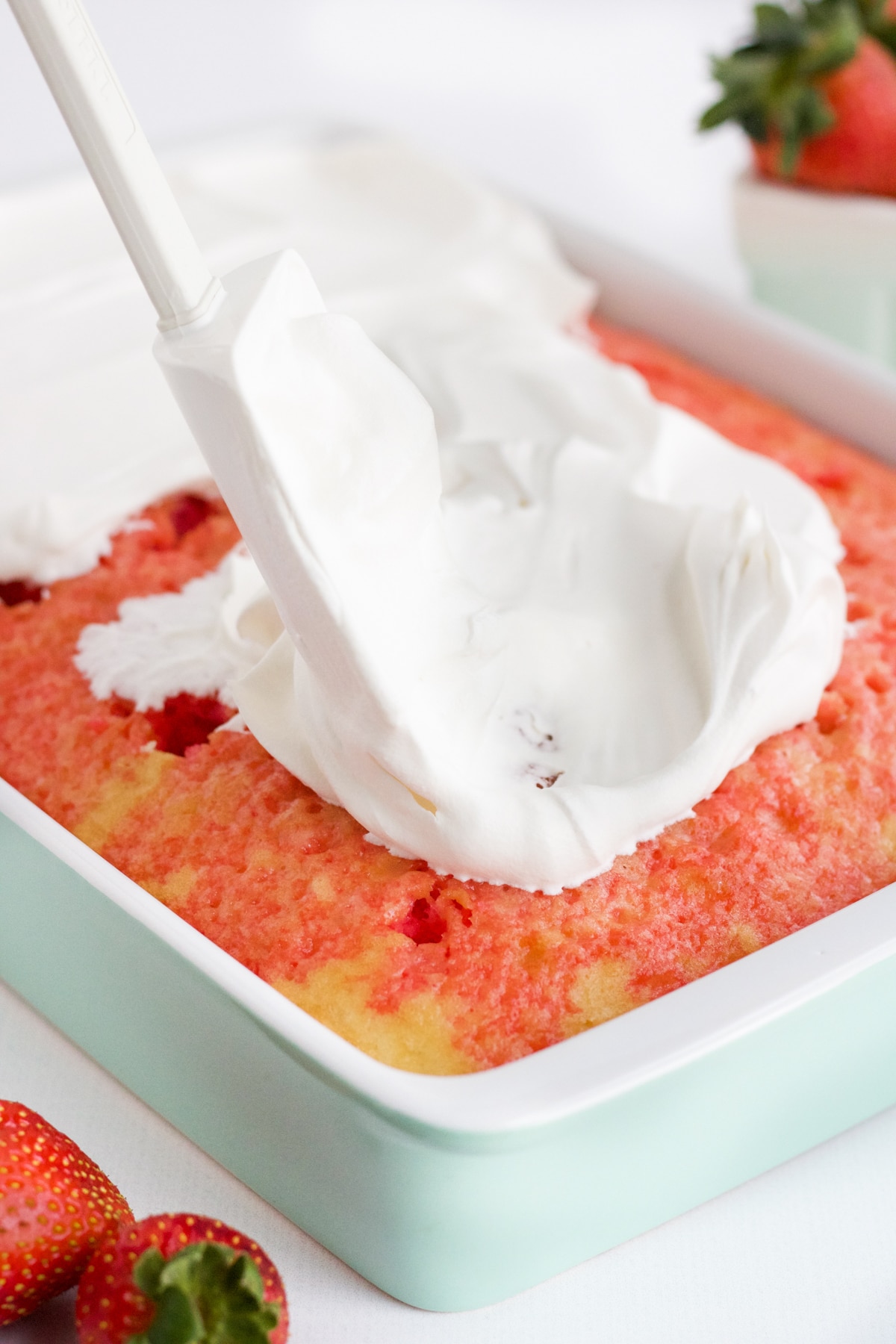 spreading whipping topping over a chilled strawberry cake in a ceramic baking pan
