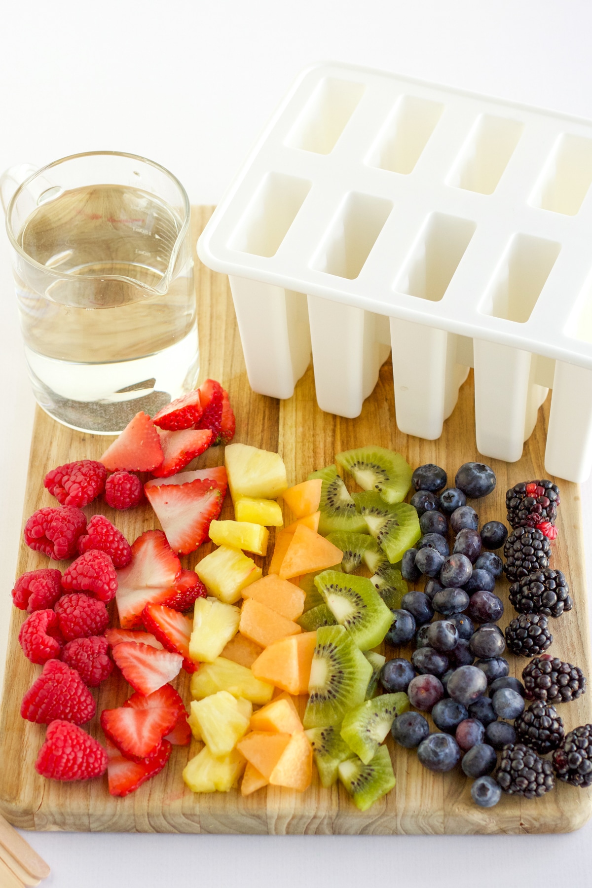 rainbow colored cut fruit on a cutting board with a glass of clear juice and a popsicle mold