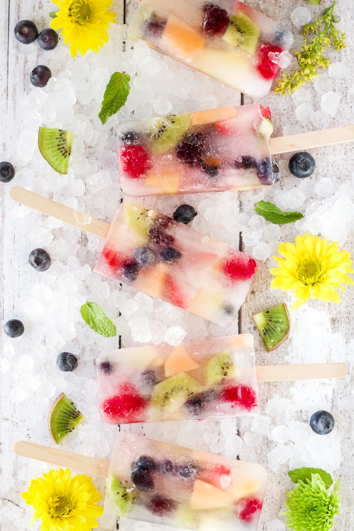 5 fruit popsicles laying on crushed ice board decorated with yellow flowers and fruit