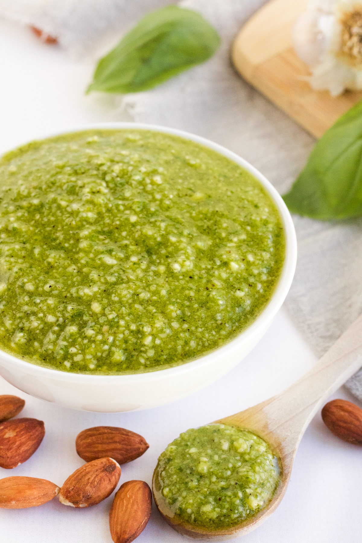 White bowl of pesto with almonds on the table with a small wooden spoon.