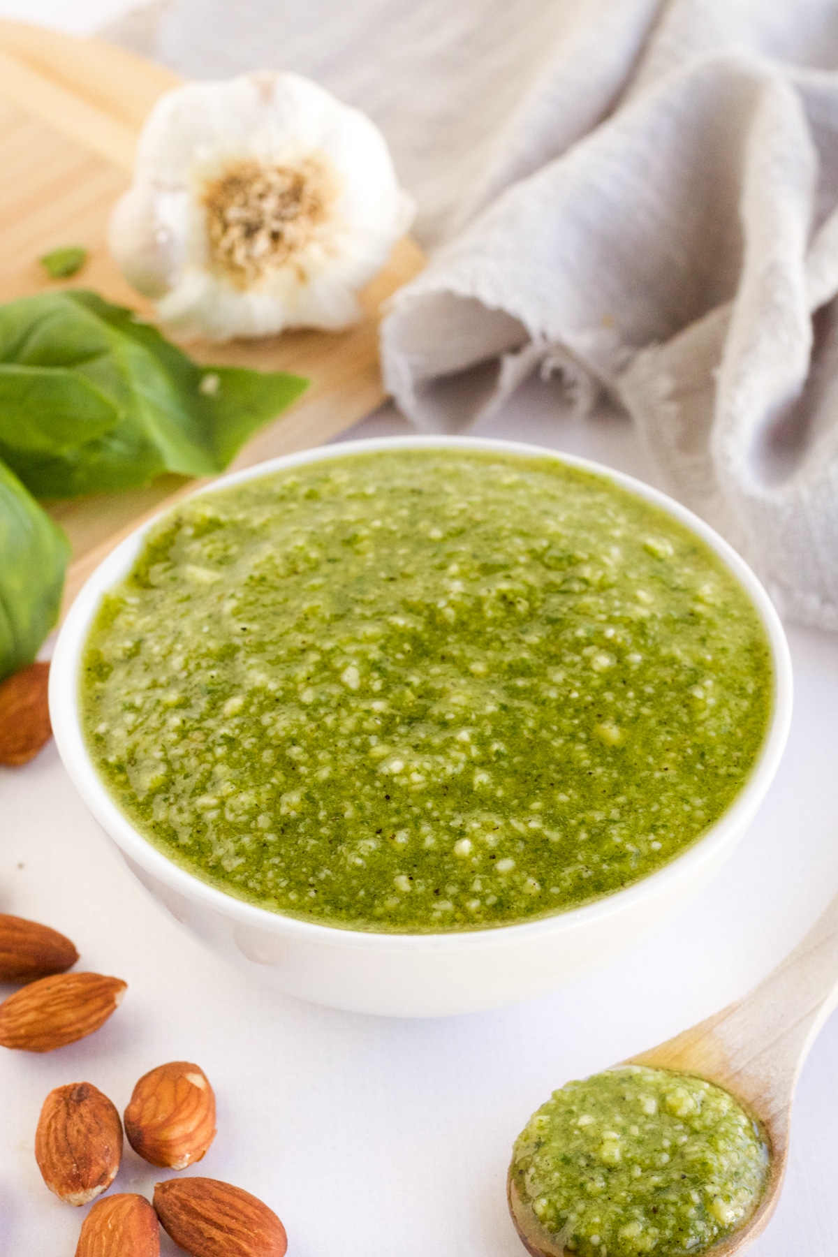 An image of homemade fresh almond basil pesto in a white bowl with garlic and basil in the background. 