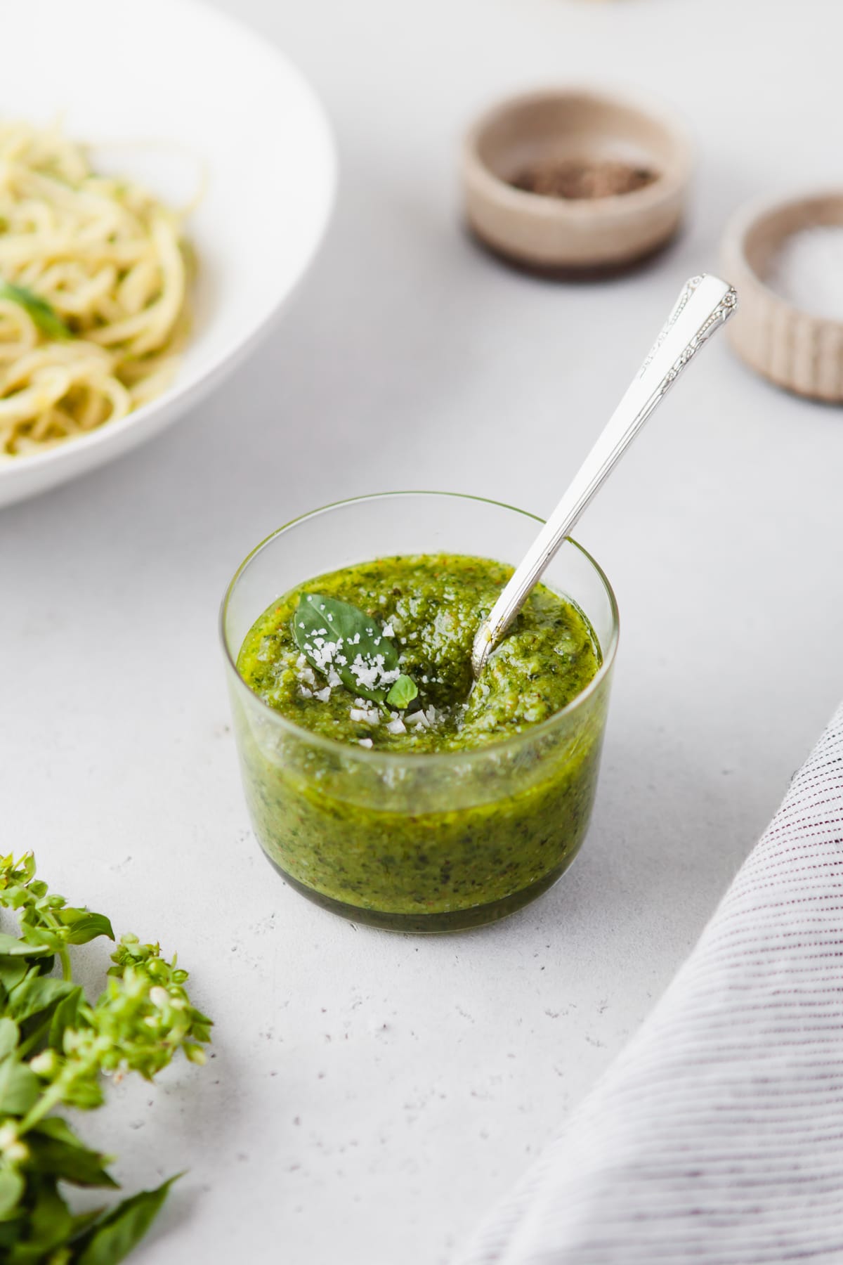 An image of cashew pesto sauce in a glass jar with pasta in the background. 