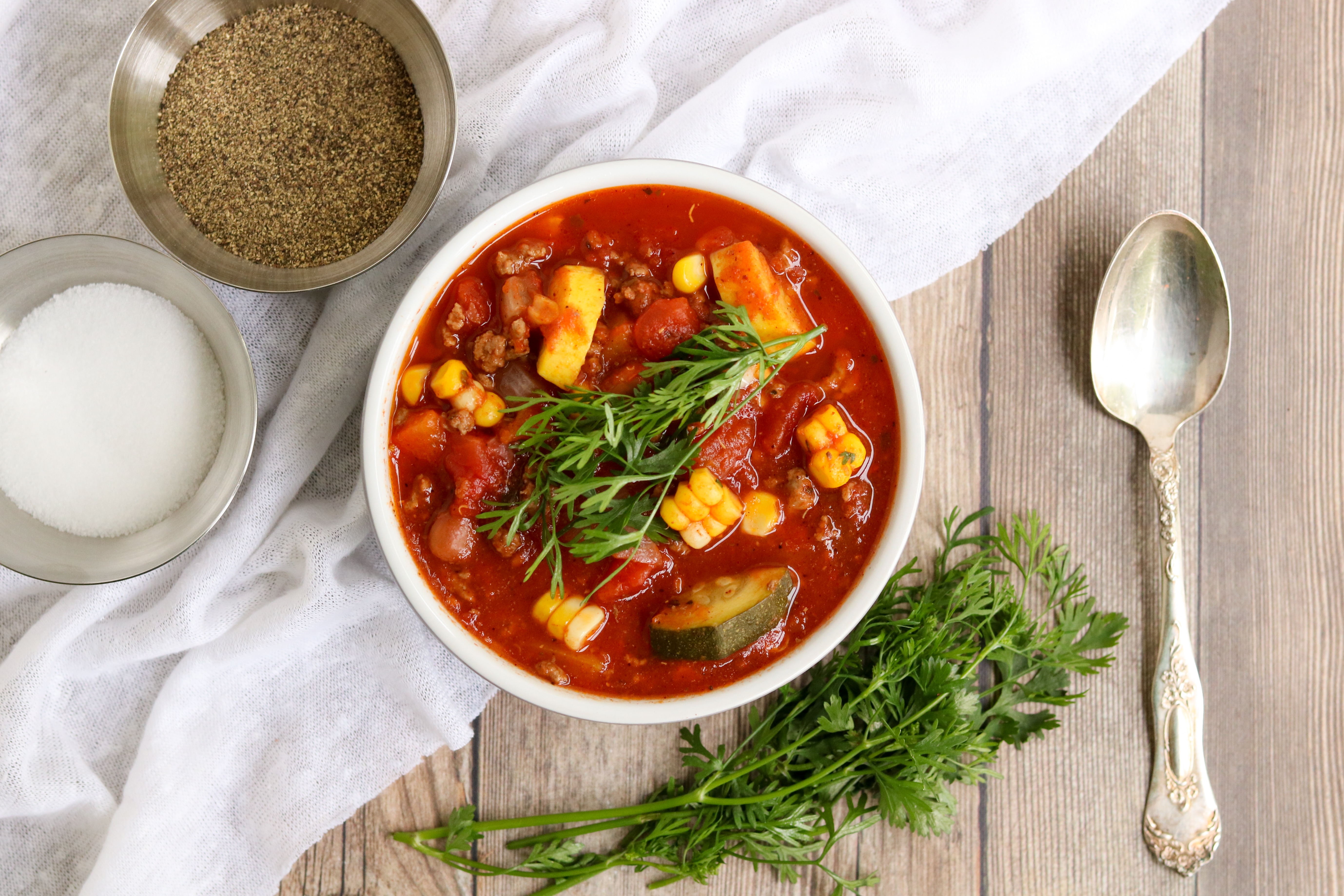 White bowl with veggie loaded chili, silver spoon, crocks of salt and pepper