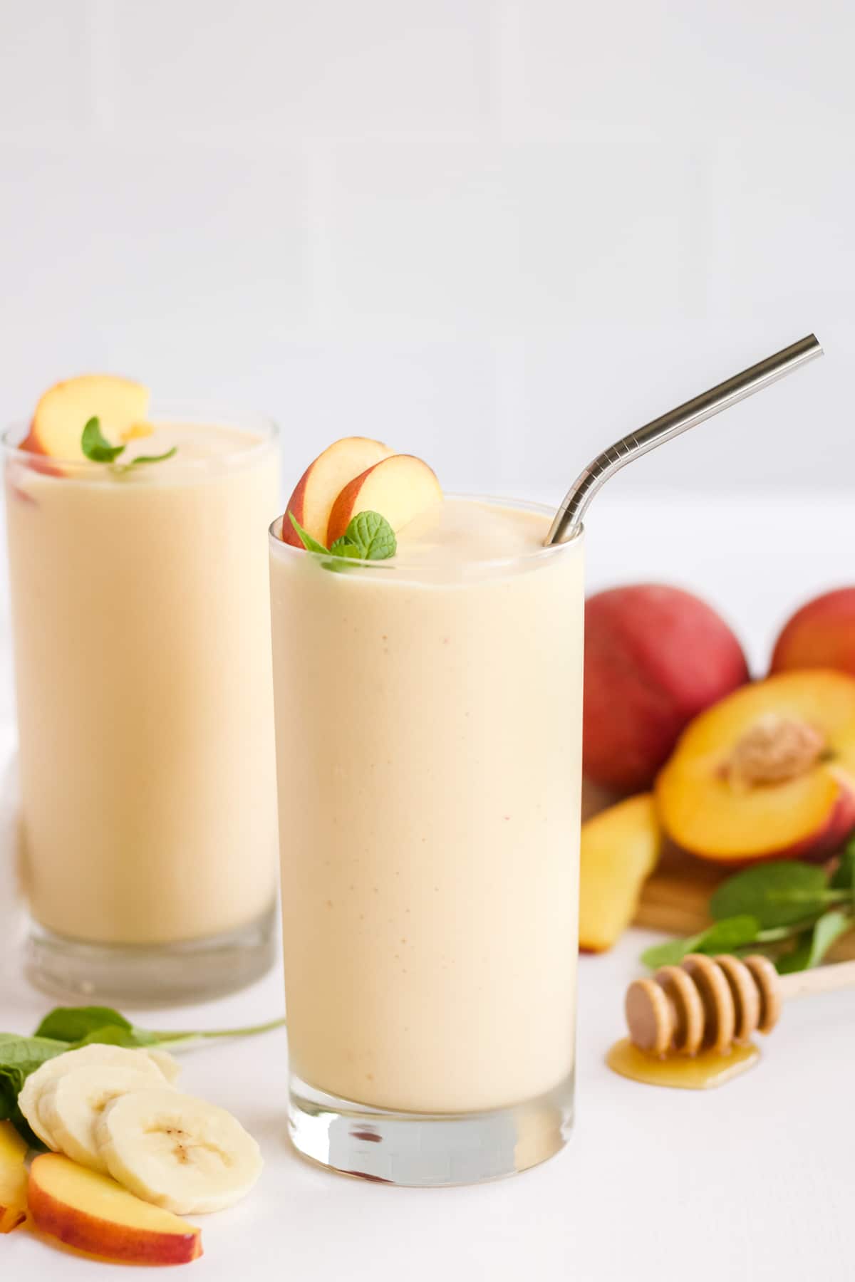 Two glasses of a peach smoothie with peaches and honey in the background.