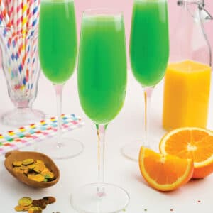 Three glasses of green mimosa on a table with coins and straws in background.
