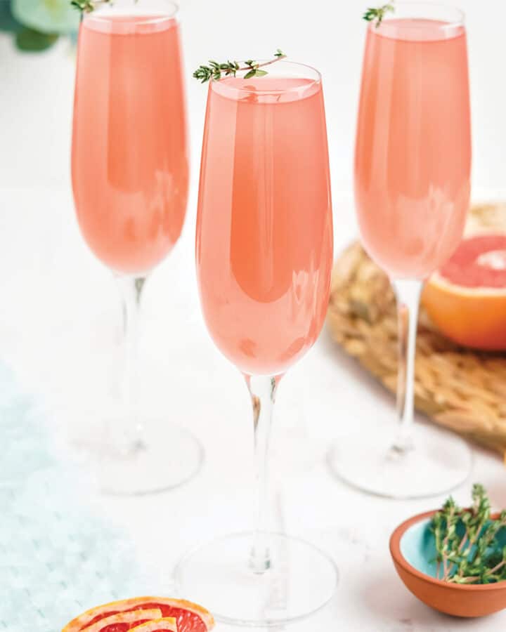 Three glasses of pink grapefruit mimosa on the table with fresh herbs on the glass as garnish.