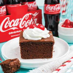 Slice of coca cola cake topped with whipped cream and a cherry.