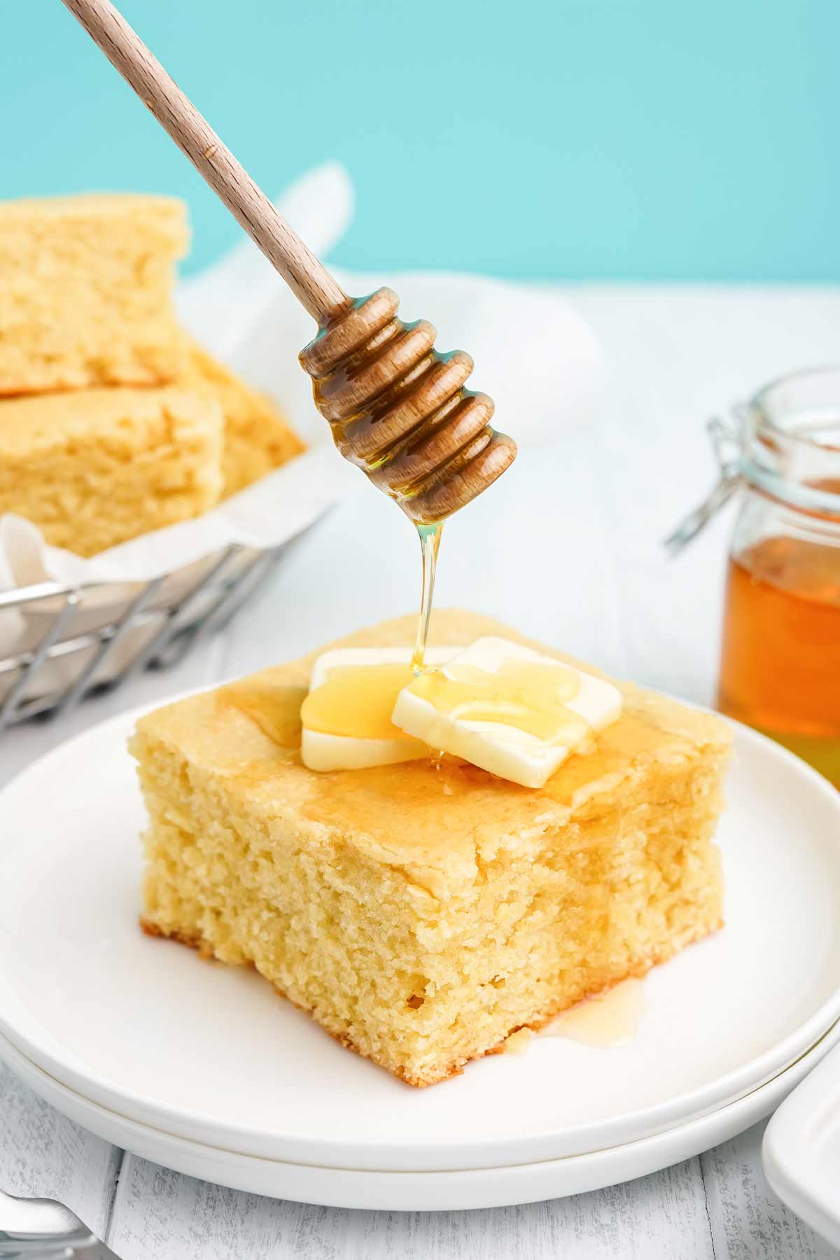 Square of cornbread with honey on a white plate with honey drizzling on top.