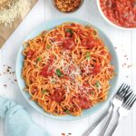Bowl of spaghetti arrabiata on the table with forks on the side, red pepper flakes and bowl of sauce.