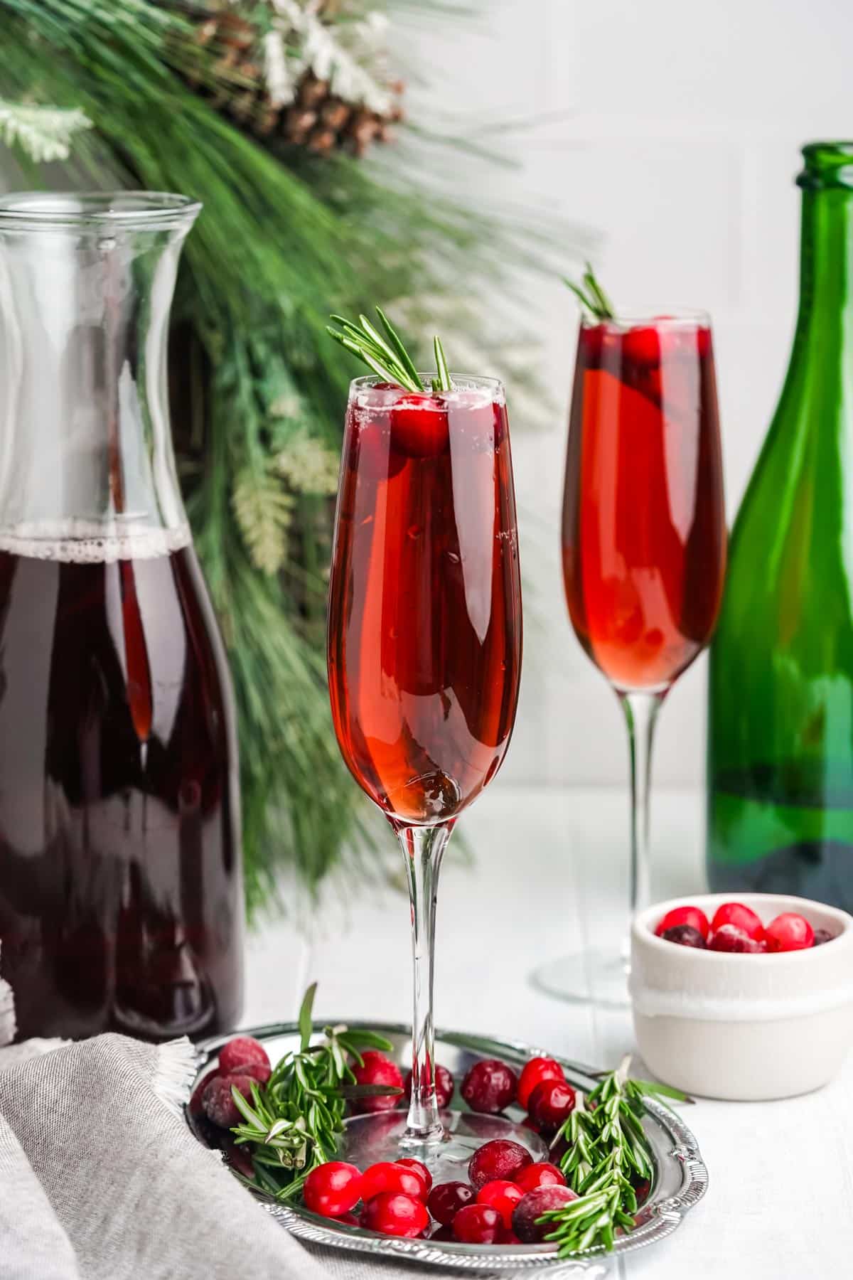 Two glasses of cranberry mimosa on the table with a bowl of fresh cranberries and carafe of juice.