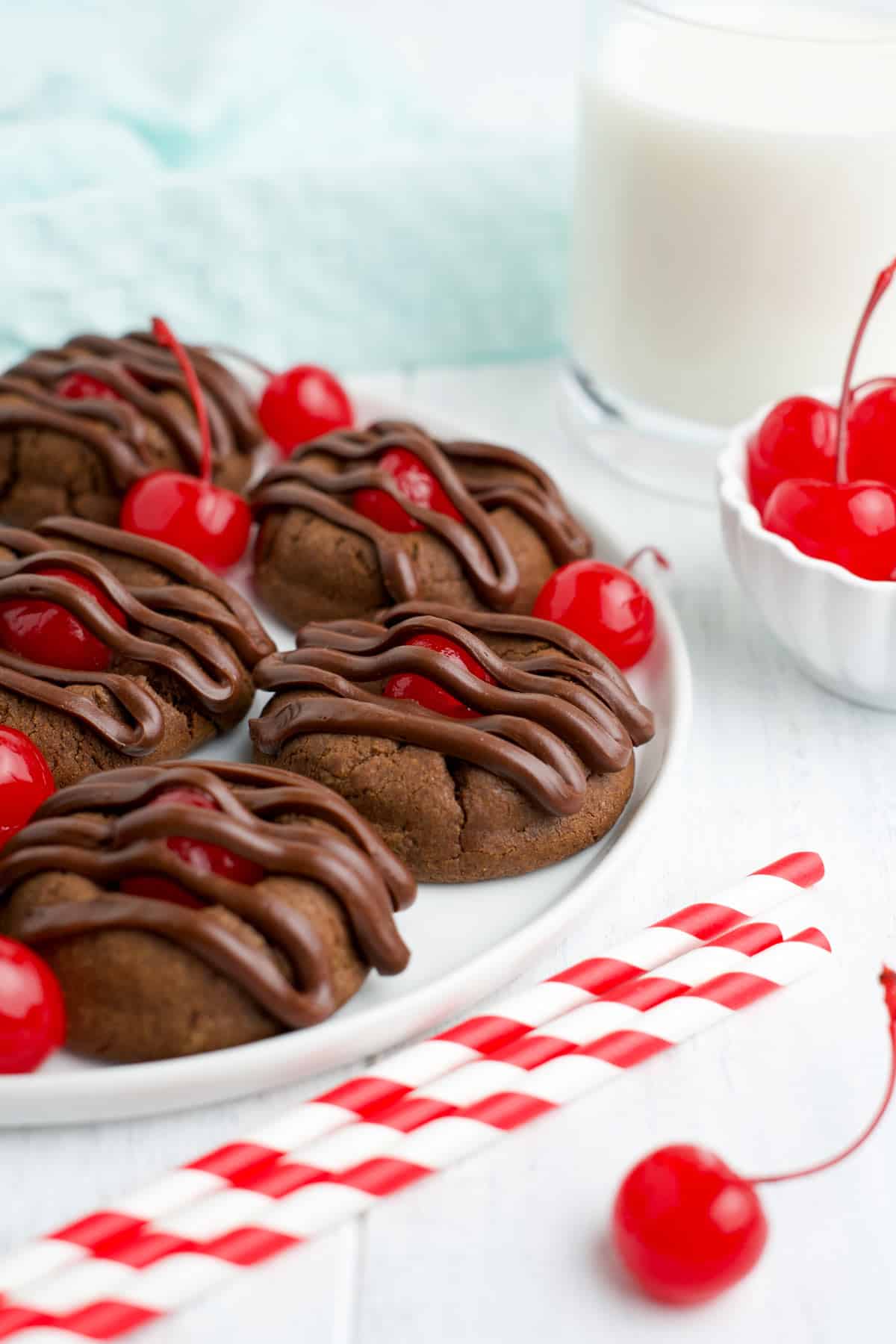 Cherry chocolate cookies on a plate with a glass of milk and a bowl of cherries.