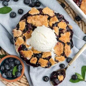 Blueberry cobbler with cake mix in a round dish with ice cream on top and fresh blueberries on the side.