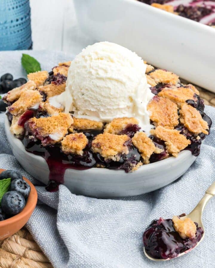 Blueberry cobbler with cake mix in a white bowl with a scoop of ice on top and a bowl of blueberries to the side.