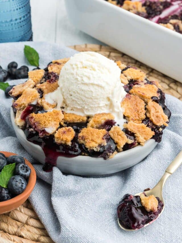 Round dish of blueberry dump cake on a blue napkin with a scoop of vanilla ice cream on top.