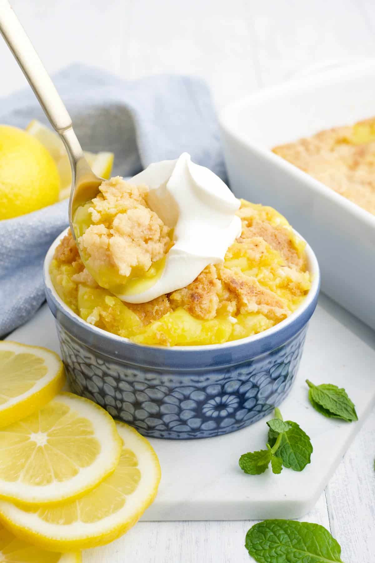 A spoon with a bite of lemon cake up over the bowl topped with whipped cream.