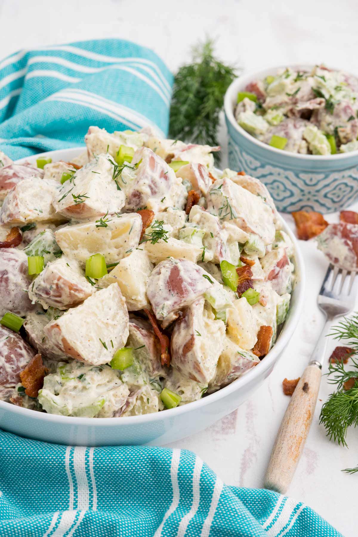 A bowl of red skin potato salad with a small bowl in the back and a fork with a potato on it.