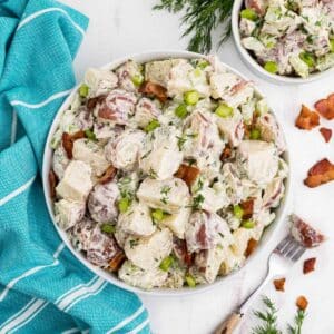 A bowl of red skin potato salad on the table with a blue tea towel and fork next to the bowl.