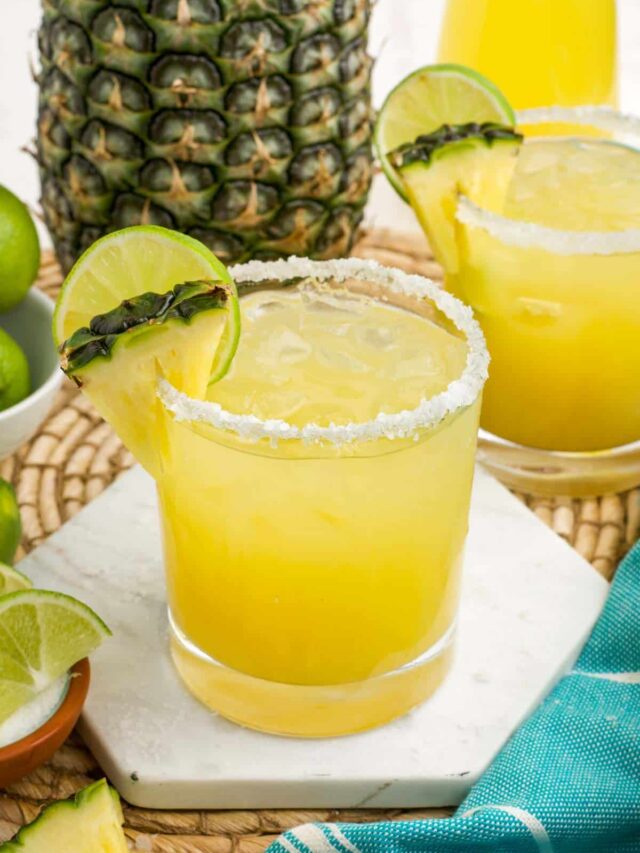 A pineapple margarita on a white coaster with fresh pineapple whole and cut on the table.