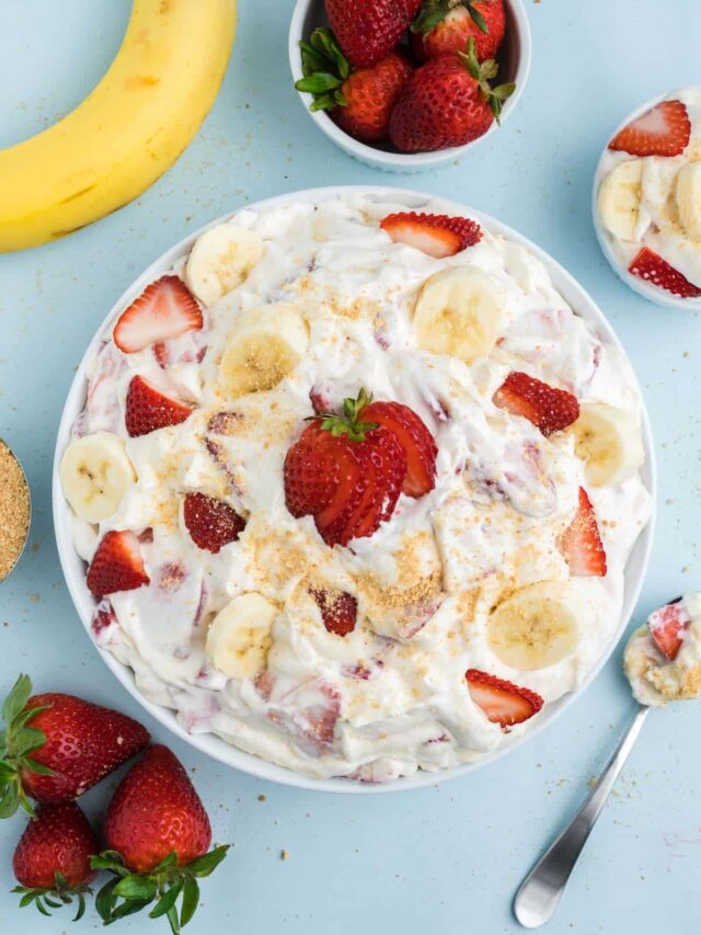 A bowl of strawberry banana cheesecake salad on the table with fresh fruit in bowls around it and a spoon to the side.